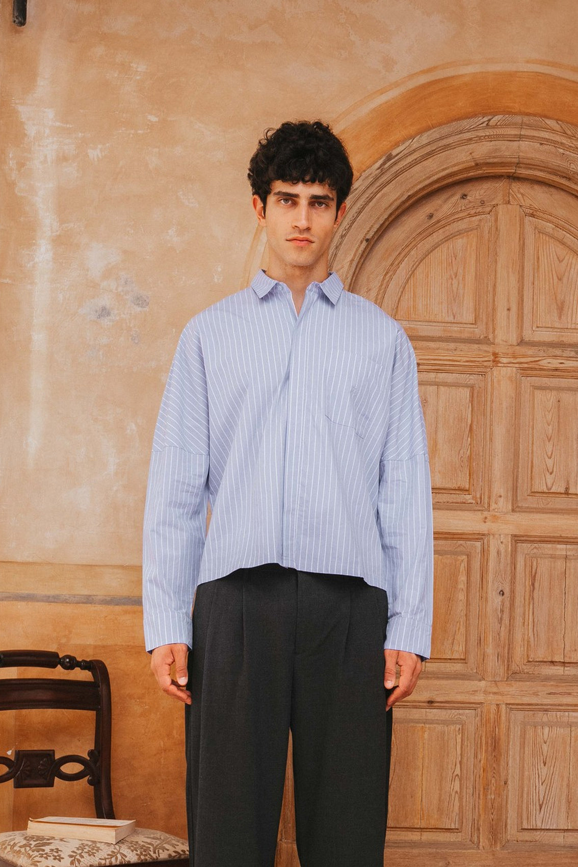 Man wearing a light blue striped shirt and dark pants standing in front of a wooden door.