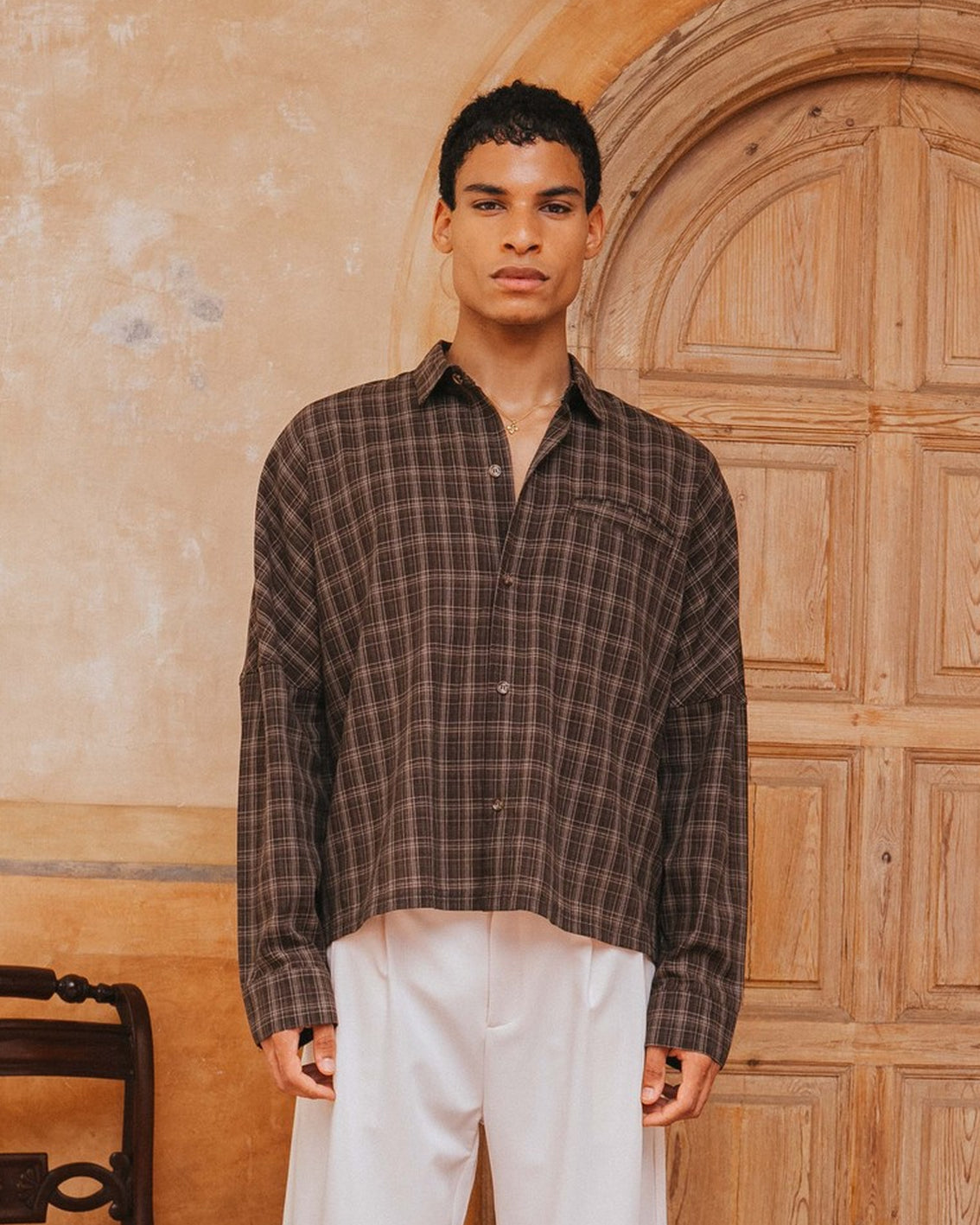 Man wearing a plaid shirt and white pants standing in front of a wooden door.